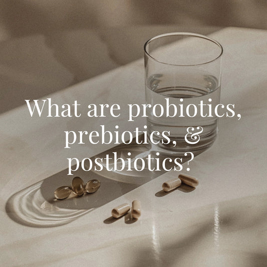 A glass of water and scattered beige supplement capsules rest on a glossy stone surface with light reflections. The scene evokes clarity and wellness. White serif text across the image reads: “What are probiotics, prebiotics, & postbiotics?”